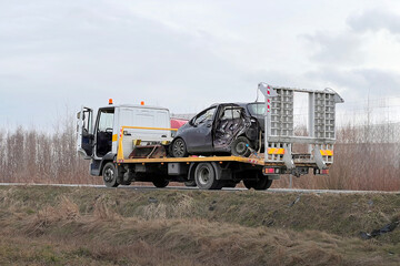 A tow truck loading a damaged black hatchback car on the highway after a collision. The car was hit on the side by another vehicle and suffered severe damage. Total loss car transported to a junkyard.