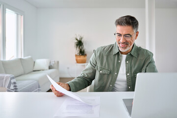Happy middle aged mature man senior entrepreneur wearing eyeglasses looking paper bill using laptop...