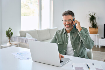 Happy middle aged mature man senior entrepreneur wearing eyeglasses talking on mobile phone looking at laptop using computer calling customer service support on smartphone working at home office.