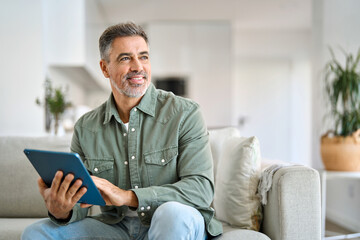 Happy middle aged man using digital tablet relaxing on couch at home. Mature male user holding tab computer holding pad technology device sitting on sofa in living room looking away. Copy space. - obrazy, fototapety, plakaty