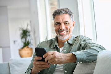 Smiling happy mature middle aged man holding cell mobile phone using smartphone sitting at home on couch, scrolling social media, checking financial apps, buying online, looking at camera. Portrait - obrazy, fototapety, plakaty