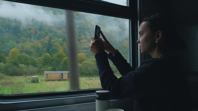 Young woman in train. Girl takes photo of mountain landscape outside the window while sitting inside transport. Passenger or traveler takes photos with phone. Woman taking pictures of Carpathian