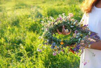 summer background. wreath of meadow flowers in girl hands outdoor, natural sunny backdrop. floral traditional decor for Summer Solstice Day, Midsummer, Litha holiday. pagan wiccan witch ritual. - obrazy, fototapety, plakaty