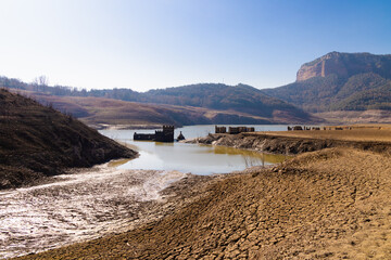 Un arroyo serpenteante fluye hacia el embalse de Sau, delineando las tierras cuarteadas y conectando con las estructuras sumergidas y los acantilados del Collsacabra en Osona, Cataluña.
