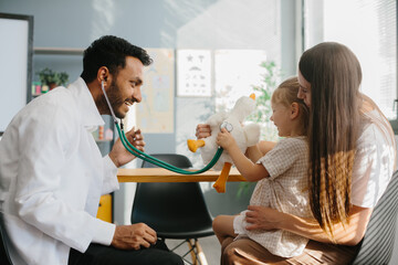 The pediatrician establishes contact, trust and a good relationship with the child. Happy little girl, mother and family doctor playing fun game with toy and stethoscope
