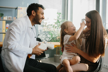 Fototapeta premium A young male pediatrician plays with a little girl patient in order to gain trust and conduct an examination. A young mother with a preschool-aged daughter in the doctor's office.