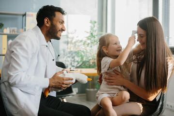 Obraz premium A young male pediatrician plays with a little girl patient in order to gain trust and conduct an examination. A young mother with a preschool-aged daughter in the doctor's office.