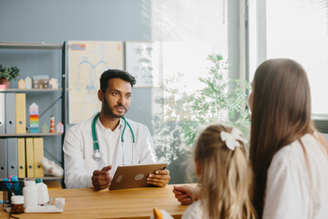 Fototapeta premium A young male pediatrician with a tablet in his hands consults a mother with her little daughter. The concept of a children's doctor appointment.