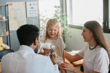 A preschool-aged girl with her mother is being examined by a pediatrician. A young man, a pulmonologist, conducts an examination using a phalendoscope. - obrazy, fototapety, plakaty
