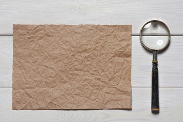 Blank crumpled paper sheet page and magnifying glass loupe on the wooden flat lay desk table background. Top view.  Secret information concept. Looking for the answers. Searching of clues concept.