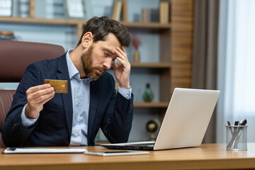 A mature adult businessman in a suit looks stressed while holding a credit card, working at his...