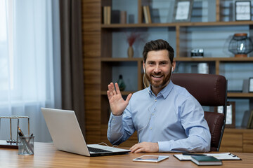 Fototapeta na wymiar Cheerful mature man in a business suit waving during a video conference in his well-organized home office environment.