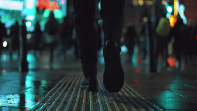 Rear back view male feet steps crossing busy street at night. Man wearing dark leather boots goes on pedestrian crossing in city. Low angle people crowd legs walking sidewalk, rainy autumn weather