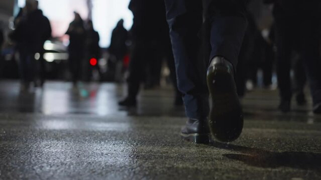 Rear back view male feet steps crossing busy street at night. Man wearing dark leather boots goes on pedestrian crossing in city. Low angle people crowd legs walking sidewalk, rainy autumn weather