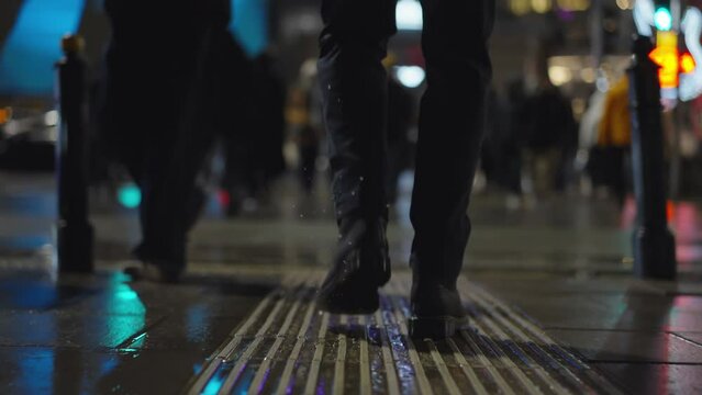 Rear back view male feet steps crossing busy street at night. Man wearing dark leather boots goes on pedestrian crossing in city. Low angle people crowd legs walking sidewalk, rainy autumn weather