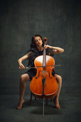 Fototapeta na wymiar Elegant, passionate musician, beautiful woman in black dress sitting and playing cello against dark green vintage background. Concept of classical art, retro style, music, inspiration, orchestra event