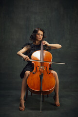 Fototapeta na wymiar Beautiful young woman, cellist sitting on chair with cello, looking at camera on vintage green background. Cultural events with classic music performances. Concept of classical art, retro style, music