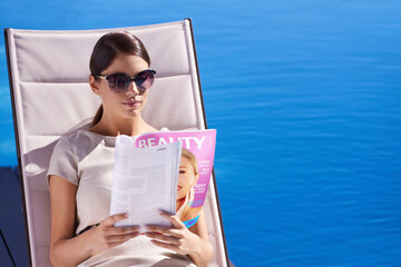 Woman, reading and magazine on sunbed by pool with sunglasses for relax, article and lifestyle...