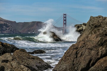 Beautiful shot of sea waves crashing on massive rocks with a background of the Golden Gate Bridge