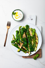 Grilled green spring vegetables: zucchini, asparagus, broccolini with yoghurt sauce on white marble background. Top view, copy space. .