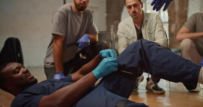 Close-up shooting: A Black male doctor in a blue medical uniform demonstrates the tightening of a medical tourniquet on his leg for public. Practical trainings on first medical aid. Course of a medic