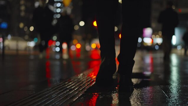 Rear back view male feet steps crossing city street at night. Man wearing dark leather boots goes on sidewalk in city. Low angle legs walking sidewalk, rainy autumn weather, reflected car lights