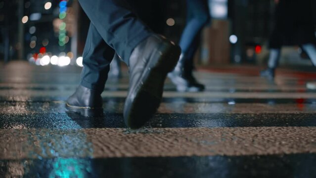 Rear back view male feet steps crossing busy street at night. Man wearing dark leather boots goes on pedestrian crossing in city. Low angle people crowd legs walking sidewalk, rainy autumn weather