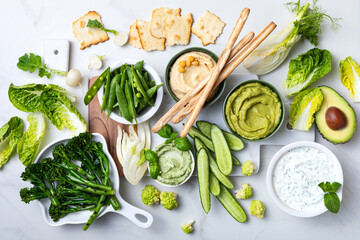 Green spring vegetable appetizer platter with dips. Healthy crudites snack board on white marble...