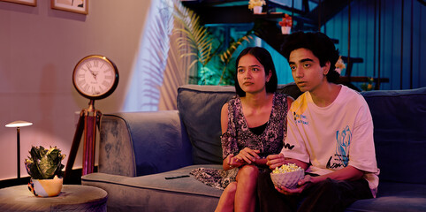 Indian Asian teen girl boy couple pair sitting sofa couch watch horror film movie TV enjoy fun joy late night indoor home gen z male female man lady friend eat snack hold bowl look scared action video