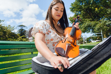 young female violinist street performer sitting outdoors preparing, taking out violin bow from case.
