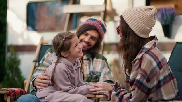 Happy little girl in a pink hoodie communicates with her mom, a brunette girl in a white hat and plaid shirt, and with her dad, a man in a green plaid shirt, during her vacation and picnic near a