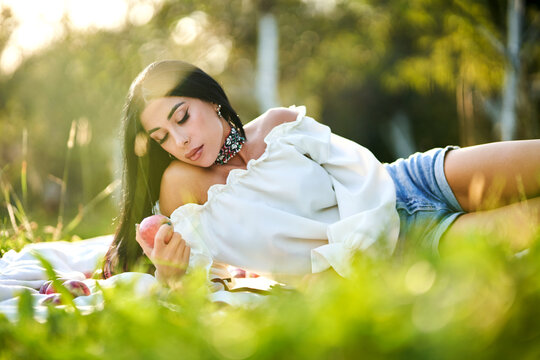 Pensive woman with harvested apple, resting on picnic rug at sunny day. Low angle view of thoughtful beautiful lady with vivid necklace, lying in green grass in apple orchard. Picnic, beauty concept. 