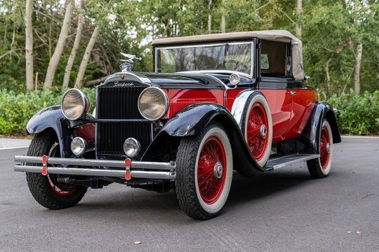 Glossy, two-tone red and black luxury Packard car parked alongside a row of lush green trees