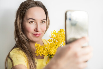 Beautiful young woman with mimosa flowers taking a selfie on white background