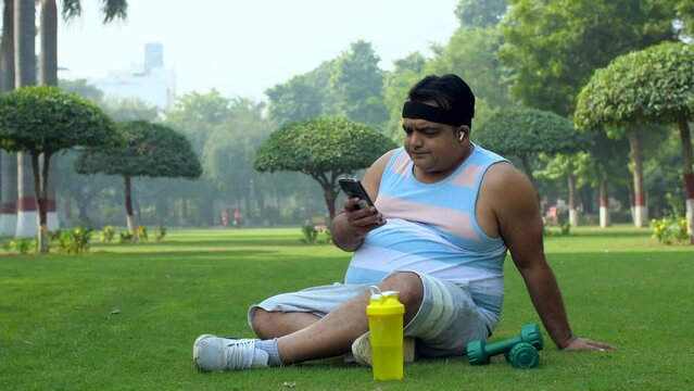 Closeup shot of a fatty Indian man listening to songs in his free time in a park -relaxing after morning walk. A tired man using phone and listening to songs while sitting in a park: post-workout r...