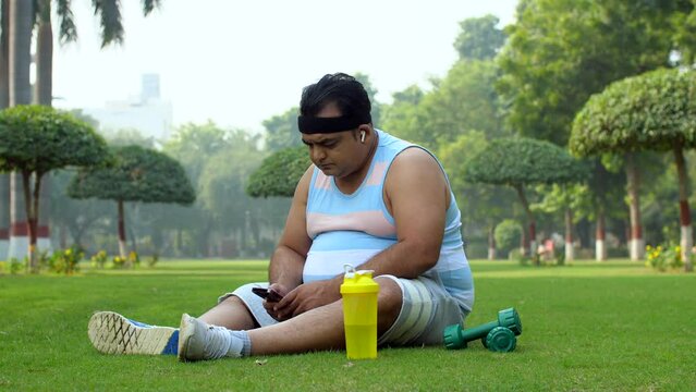 Middle-aged man using smartphone in free time post workout - time pass  fat man  losing weight. Indian man scrolling through his mobile phone after tiring fitness session - wireless earphones  fat ...
