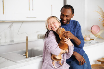 Joyful multiethnic couple in sweaters playing with chihuahua dog in modern kitchen at home