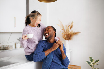 Laughing asian woman holding coffee and hugging african american boyfriend in kitchen