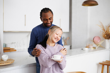 Smiling young african american man in sweater hugging asian girlfriend with coffee in kitchen in...