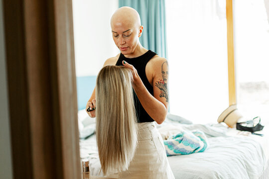 Bald woman with cancer holding blond wig standing in bedroom at home