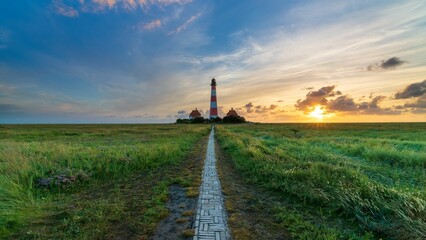 Scenic pathway through a lush grassy field leading to a lighthouse in the distance at sunset.