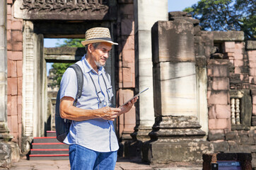 senior man wearing hat with backpack, camera and tablet computer in hand is searching data at...