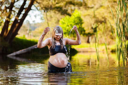 Pregnant Aboriginal woman in traditional body paint splashing streams of water droplets in creek