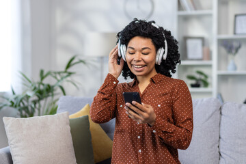 Close-up photo of a smiling young African-American woman sitting on the sofa at home, wearing headphones and using a mobile phone