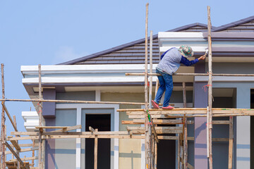 Painter on wooden scaffolding structure is Painting roof eaves of modern House against blue sky...
