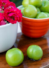 Front view of two, green plums lying on wet wooden board, table, bowls with pink roses and plums on background. Concept of summer.