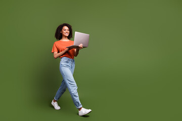 Full length photo of pretty cute woman wear orange t-shirt texting modern gadget walking empty space isolated green color background