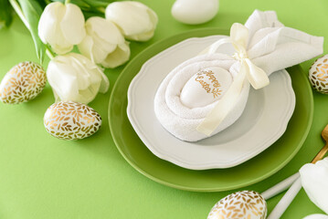 Obraz na płótnie Canvas Plate decorated bunny ear napkin, white tulips and Easter eggs on green background. Easter celebration concept. Flat lay. Top view