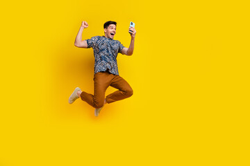 Fototapeta na wymiar Full size photo of overjoyed guy wear stylish shirt brown pants look at smartphine jumping win lottery isolated on yellow color background