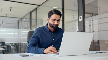 Smiling Indian business man looking at laptop using computer working in office. Busy professional businessman employee or company executive searching online solutions at workplace. - Powered by Adobe